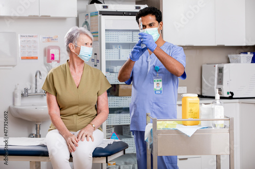 A Young Indian Male Medical Nurse Administering a Covid-19 Vaccine with a Syringe Needle to an Elderly Senior Female Patient Wearing Generic ID Badge, Gloves and Mask in Hospital or Health Clinic.
