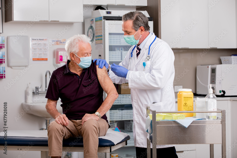 A White Male Medical Doctor Administering a Covid-19 Vaccine Injection with a Syringe Needle to an Elderly Senior Male Patient Wearing Generic ID Badge, Gloves and Mask in Hospital or Health Clinic. A White Male Medical Doctor Administering a Covid-19 Vaccine Injection with a Syringe Needle to an Elderly Senior Male Patient Wearing Generic ID Badge, Gloves and Mask in Hospital or Health Clinic.