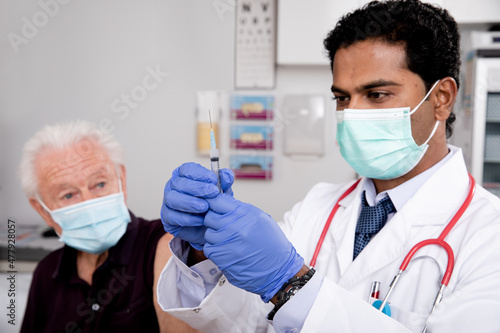 A Young Indian Male Medical Doctor Administering a Covid-19 Vaccine with a Syringe Needle to an Elderly Senior Male Patient Wearing Generic ID Badge, Gloves and Mask in Hospital or Health Clinic.