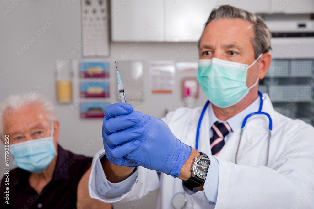 A White Male Medical Doctor Administering a Covid-19 Vaccine Injection with a Syringe Needle to an Elderly Senior Male Patient Wearing Generic ID Badge, Gloves and Mask in Hospital or Health Clinic. A White Male Medical Doctor Administering a Covid-19 Vaccine Injection with a Syringe Needle to an Elderly Senior Male Patient Wearing Generic ID Badge, Gloves and Mask in Hospital or Health Clinic.