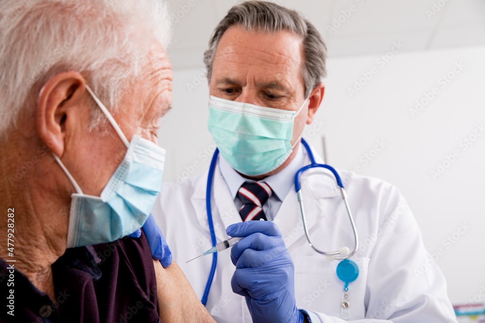 Fototapeta premium A White Male Medical Doctor Administering a Covid-19 Vaccine Injection with a Syringe Needle to an Elderly Senior Male Patient Wearing Generic ID Badge, Gloves and Mask in Hospital or Health Clinic.