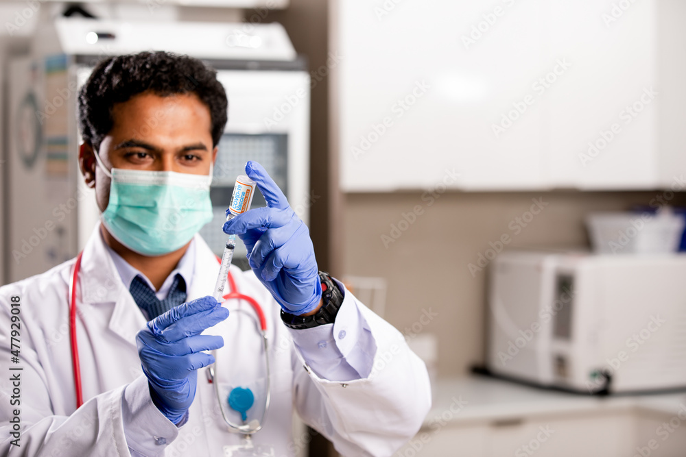 A Young Indian Male Medical Doctor Extracting a Covid-19 Vaccine Injection with a Syringe Needle Wearing Generic ID, Gloves, Mask, White Lab Coat and Stethoscope in Hospital or Health Clinic. - obrazy, fototapety, plakaty 