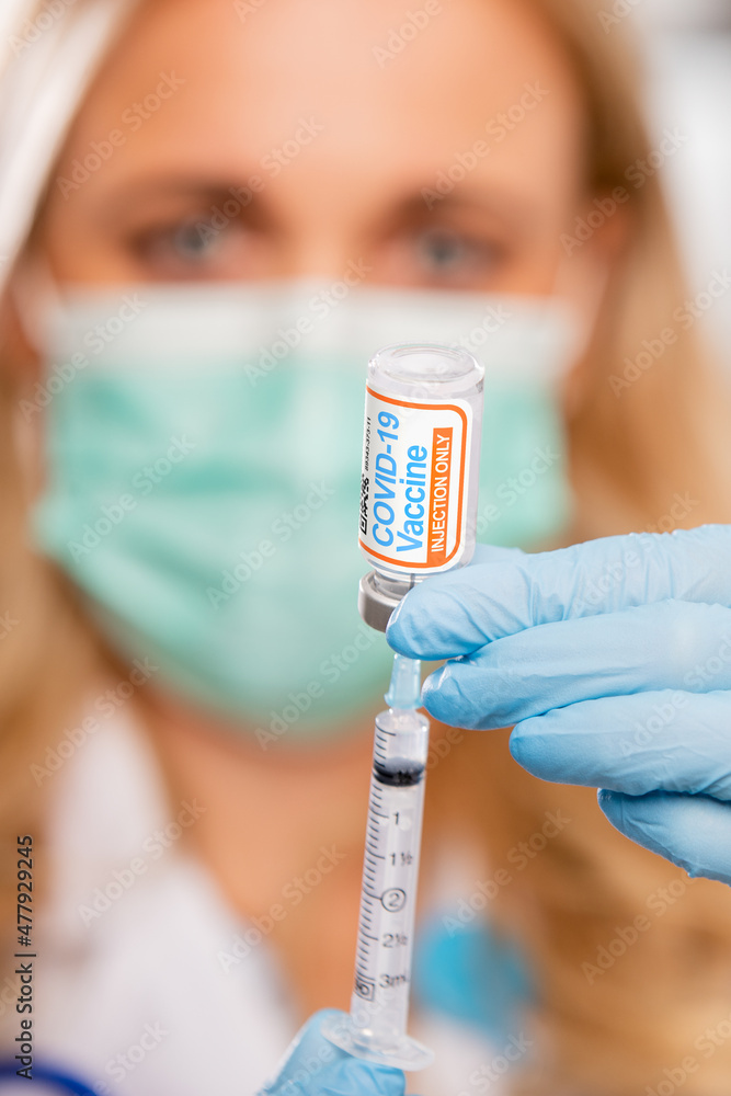 A Young Female Medical Doctor Extracting a Covid-19 Vaccine with a Syringe Needle Wearing Generic ID Badge, Gloves, Mask, White Lab Coat and Stethoscope in Hospital or Health Clinic. A Young Female Medical Doctor Extracting a Covid-19 Vaccine with a Syringe Needle Wearing Generic ID Badge, Gloves, Mask, White Lab Coat and Stethoscope in Hospital or Health Clinic.