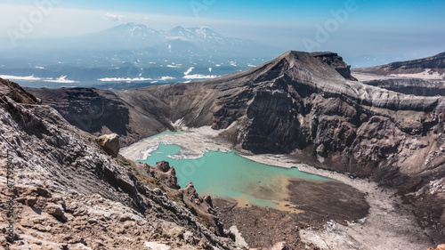 Turquoise acid lake in the crater of the Gorely volcano. Deposits of snow and sulfur on the shores. The layered structure of steep slopes. In the distance, against the blue sky - mountains photo