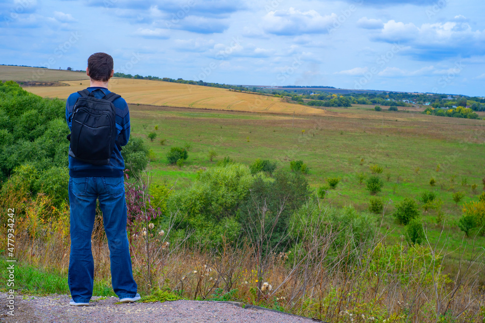 Back of a man tourist with a backpack on the background of a beautiful field and hills Back of a man tourist with a backpack on the background of a beautiful field and hills