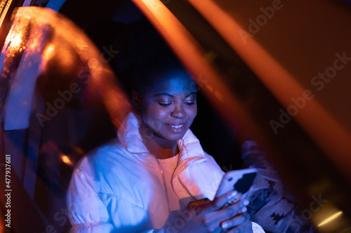 African woman suffer from gadget addiction spend time sitting in darkness in car checking social media, texting and scrolling mobile web applications. Young black female use smartphone inside vehicle