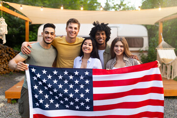 Diverse millennial friends holding American flag and smiling at camera in front of motorhome at campsite