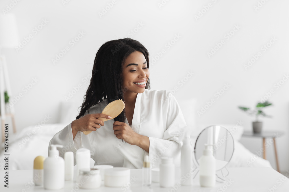 Happy young black lady in bathrobe sitting in bedroom, combing her curly hair with wooden brush, copy space