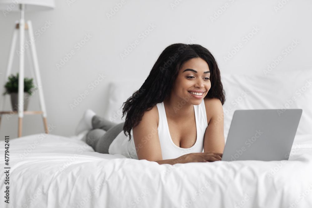 Job in bedroom, browsing on internet. Excited young african american woman working on laptop, lying on bed Job in bedroom, browsing on internet. Excited young african american woman working on laptop, lying on bed