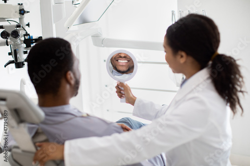 Black Guy Looking At Mirror, Checking His Teeth After Dental Treatment