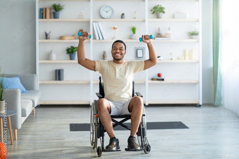 Fototapeta premium Rehab exercises for disabled people. Paraplegic black guy in wheelchair working out with dumbbells at home, full length