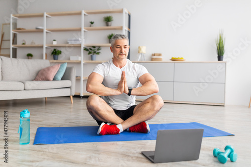 Mature man sitting in lotus pose in living room interior in front of laptop, meditating, working out at home, copy space