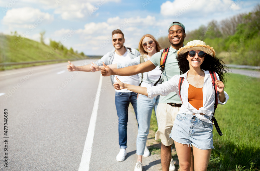 Young multiracial friends standing on highway, showing hitchhiking gesture, stopping car, going on journey together