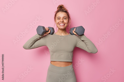 Horizontal shot of satisfied motivated sportswoman has regular training raises arms with dumbbells does exercises for muscles dressed in sportswear smiles toothily isolated on pink wall Fotobehang