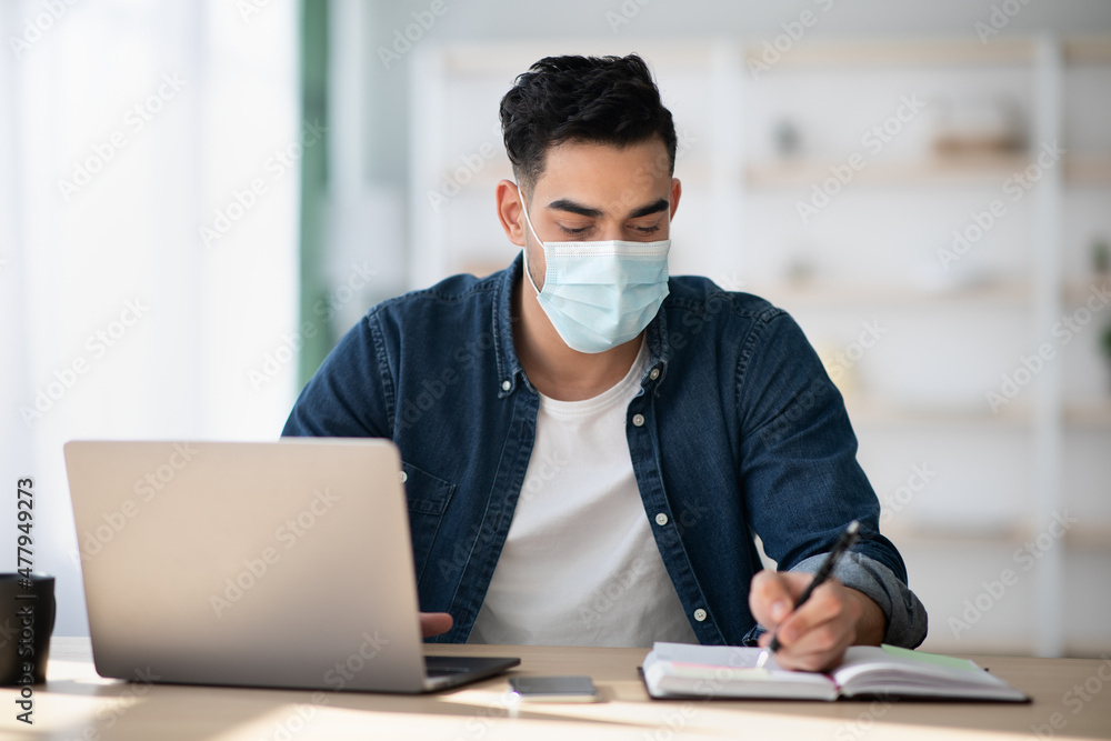 Arab man in protective face mask using laptop, taking notes