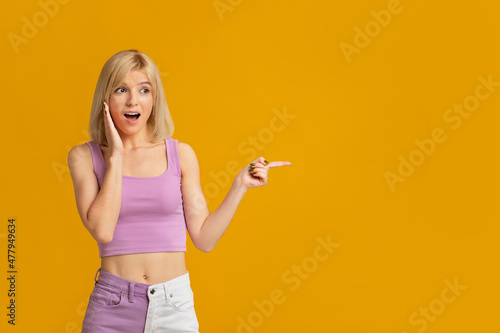 Wow, look there. Shocked young lady pointing finger at empty space standing on yellow background, studo shot photo