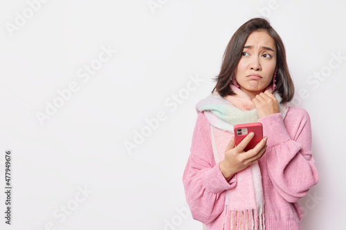 Unhappy brunette young Asian woman browses boring website on smartphone reads repetitious news holds mobile phone wears sweater and scarf around neck isolated over white background copy space