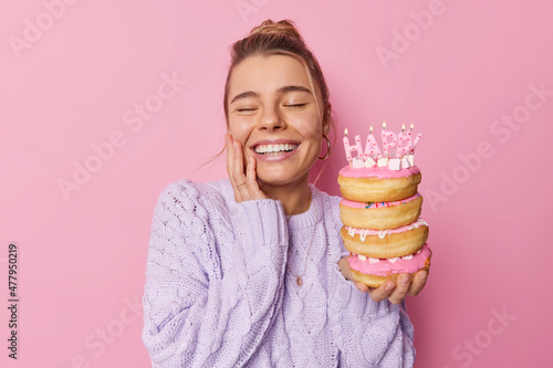 Optimistic European woman smiles broadly closes eyes from happiness celebrates special occasion holds pile of asty doughnuts dressed in knitted jumper isolated over pink background. Happy birthday photo