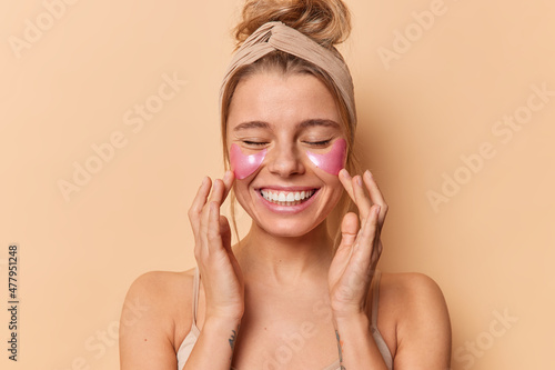 Portrait of cheerful tender woman touches face enjoys softness of skin applies hydrogel patches under eyes for reducing puffiness wears headband stands bare shoulders against beige background