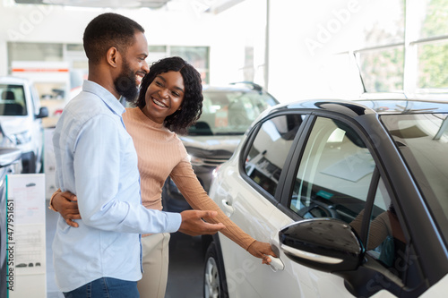Car Choosing. Happy Black Spouses Standing Next To Modern Vehicle In Showroom