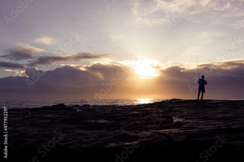 silhouette of a person standing and fishing on a rock at sunset beside the ocean
