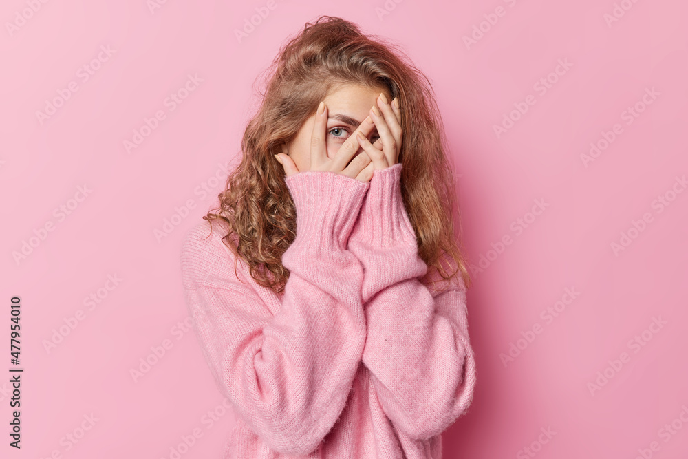 Serious long haired beautiful woman peeks through fingers covers face tries to hide looks attentively at something dressed casual jumper isolated over pink background. Horizontal studio shot Serious long haired beautiful woman peeks through fingers covers face tries to hide looks attentively at something dressed casual jumper isolated over pink background. Horizontal studio shot