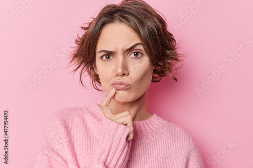 Serious skeptical woman keeps index finger near folded lips raises eyebrows suspiciously has doubts suspects someone tries to search solution wears casual jumper isolated over pink background