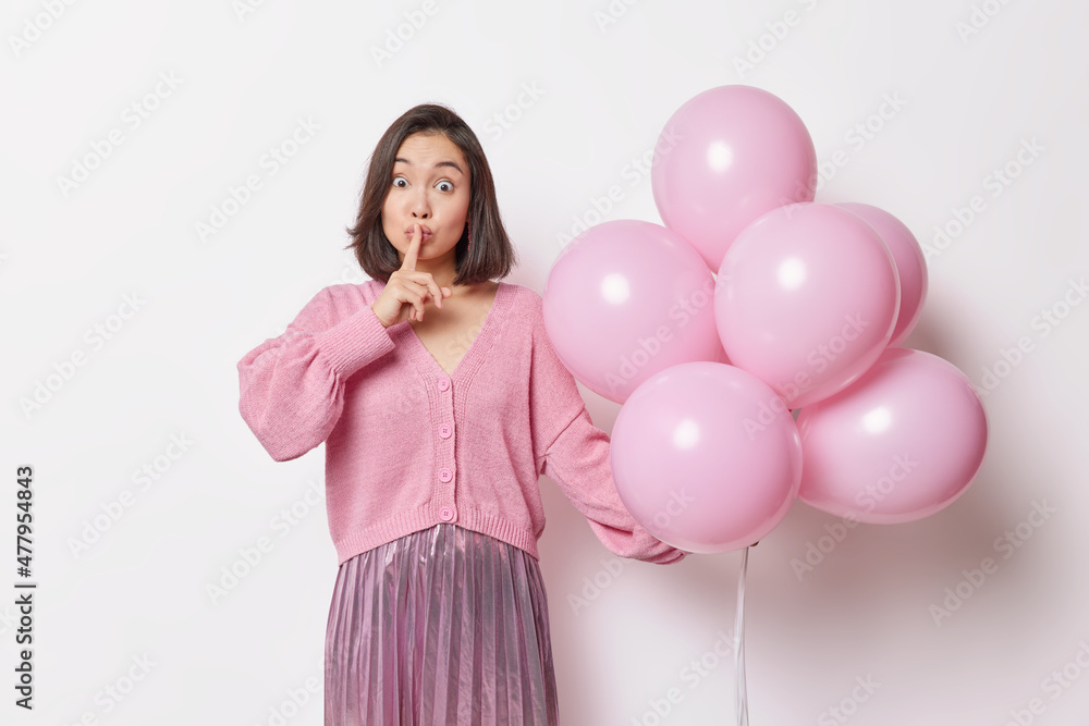 Surprised Asian woman in festive clothes makes silence gesture holds bunch of inflated balloons comes on birthday party isolated over white background. People holidays and celebration concept Surprised Asian woman in festive clothes makes silence gesture holds bunch of inflated balloons comes on birthday party isolated over white background. People holidays and celebration concept