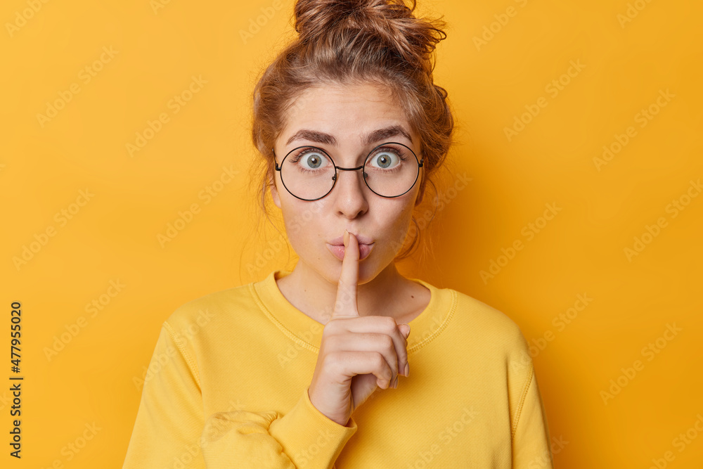 Stunned emotional beautiful woman hushing with index finger shares secret makes taboo gesture stares through round spectacles wears casual jumper isolated over yellow background. Shh be quiet - obrazy, fototapety, plakaty 