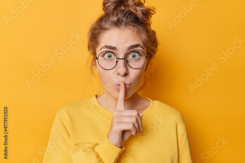 Stunned emotional beautiful woman hushing with index finger shares secret makes taboo gesture stares through round spectacles wears casual jumper isolated over yellow background. Shh be quiet photo
