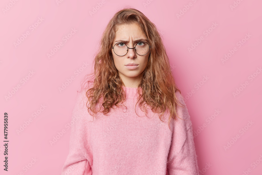 Horizontal shot of angry disappointed European woman looks unhappily at camera has grumpy expression hears something unpleasant wears round spectacles and jumper isolated over pink backgroud Horizontal shot of angry disappointed European woman looks unhappily at camera has grumpy expression hears something unpleasant wears round spectacles and jumper isolated over pink backgroud