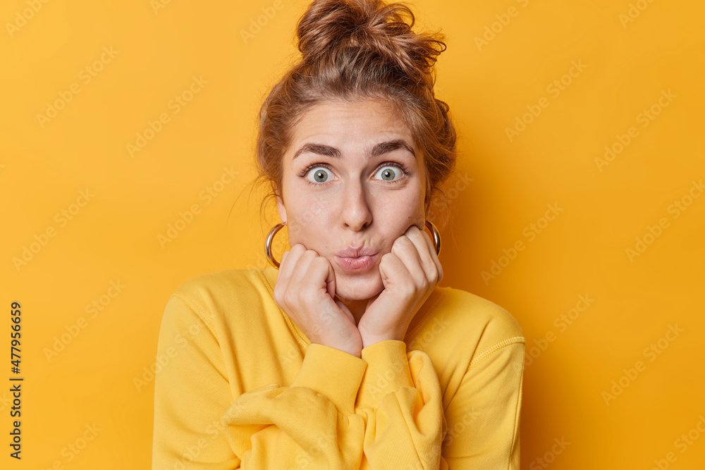 Pretty impressed woman keeps lips folded stares with widely opened eyes keeps hands under chin reacts on something exciting dressed casually isolated over vivid yellow background. Face expressions Pretty impressed woman keeps lips folded stares with widely opened eyes keeps hands under chin reacts on something exciting dressed casually isolated over vivid yellow background. Face expressions