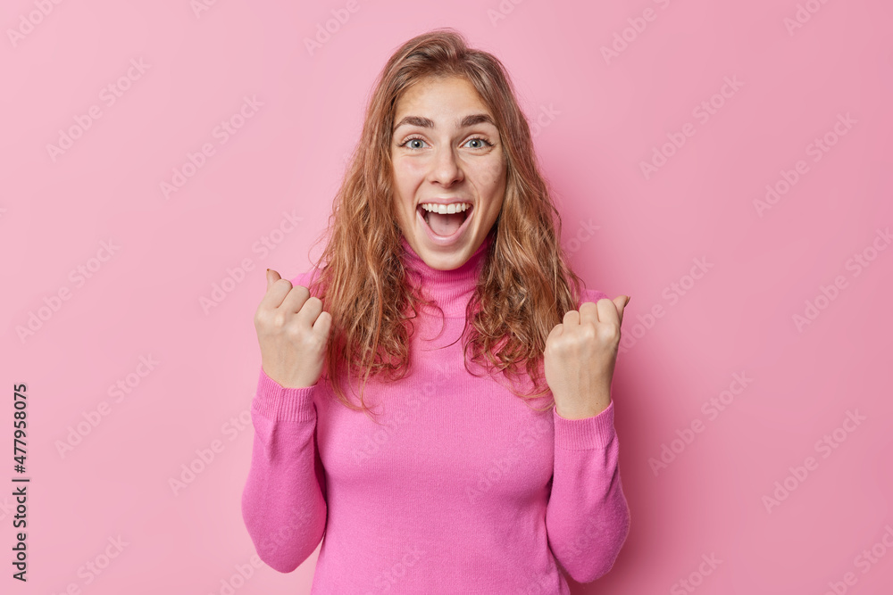 Excited joyful European woman exclaims yes clenches fists triumphs over something celebrates success wears turtleneck isolated over pink background being supportive and happy about something Excited joyful European woman exclaims yes clenches fists triumphs over something celebrates success wears turtleneck isolated over pink background being supportive and happy about something
