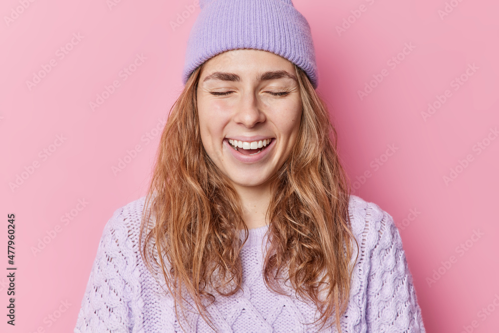Indoor shot of happy woman daydreams and closes eyes has happy memory giggles positively wears hat and knitted sweater smiles pleased poses against pink background. Positive emotions concept Indoor shot of happy woman daydreams and closes eyes has happy memory giggles positively wears hat and knitted sweater smiles pleased poses against pink background. Positive emotions concept
