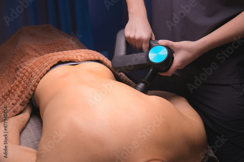 Close-up of a professional male masseur stimulates the back muscles of a male patient in a dark spa room for massage. Percussion mechanical effect on overstrained muscles