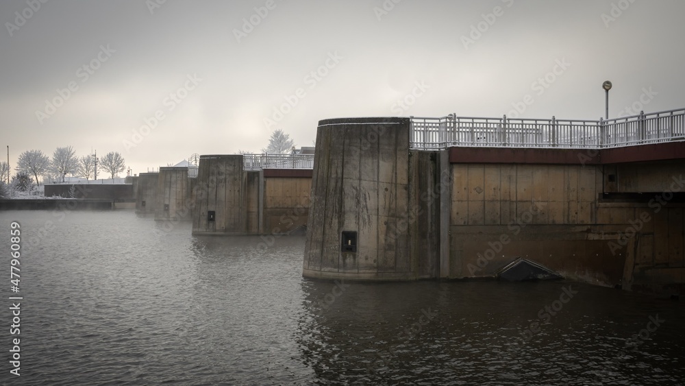 Fototapeta premium Bremen, Germany. First hoarfrost at Weserwehr weir next to swb Stadtwerke power plant beneath Weser river in foggy ice cold Hastedt. Chemical industry and moody bridges
