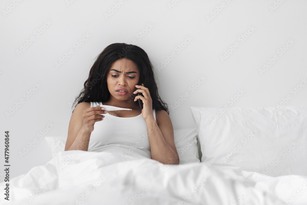 Worried ill african american woman sitting in bed, holding thermometer and calling family doctor for counseling Worried ill african american woman sitting in bed, holding thermometer and calling family doctor for counseling