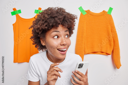 Indoor shot of happy surprised woman with curly hair looks wondered aside holds mobile phone for chatting online poses against white background with orange plastered clothes receieves great offer