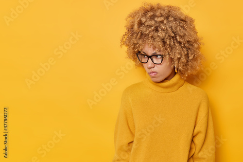 Curly haired young cute woman looks angrily away being dissatisfied with something purses lips concentrated on left wears eyeglasses and jumper isolated over yellow background with copy space © Wayhome Studio