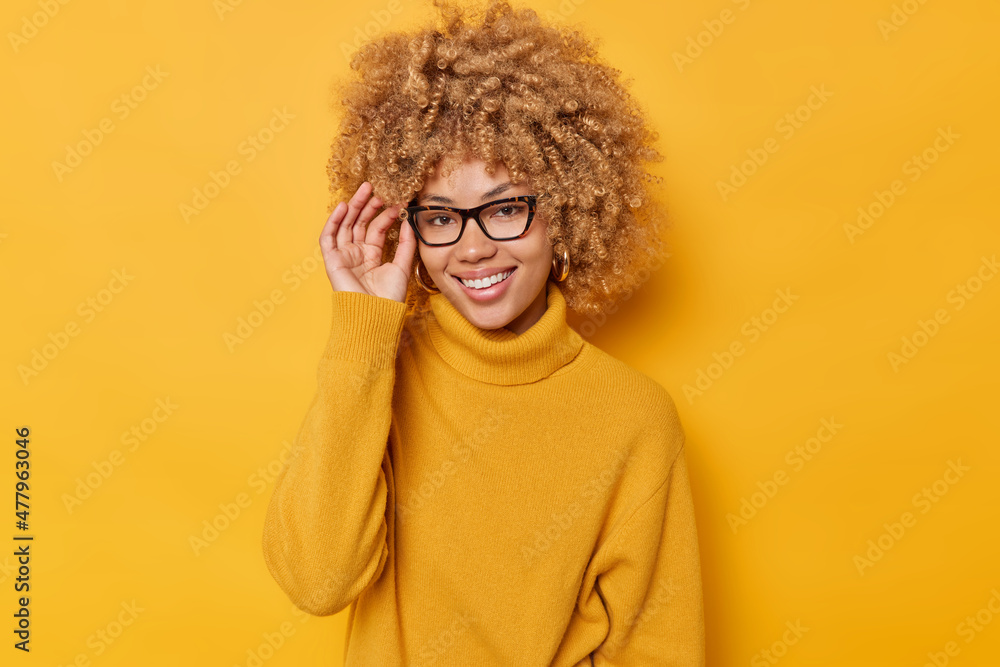 Portrait of cheerful woman keeps hand on rim of spectacles smiles joyfully wears casual jumper feels positive isolated over vivid yellow background stands pleased indoor. People and emotions Portrait of cheerful woman keeps hand on rim of spectacles smiles joyfully wears casual jumper feels positive isolated over vivid yellow background stands pleased indoor. People and emotions