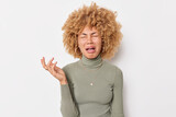 Upset crying curly haired young woman feels frustrated stands distressed keeps hand raised up wears turtleneck isolated over white background. Emotional burnoutdepression and lockdown concept.