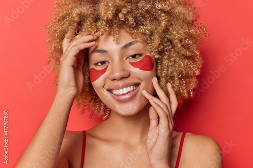 Photo of positive woman with curly blonde hair applies collagen patches under eyes for reducing wrinkles and fine lines smiles toothily isolated over red background. Skin care and wellness concept