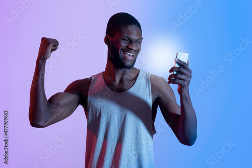 Excited black guy with smartphone making YES gesture, winning sports bet in neon light