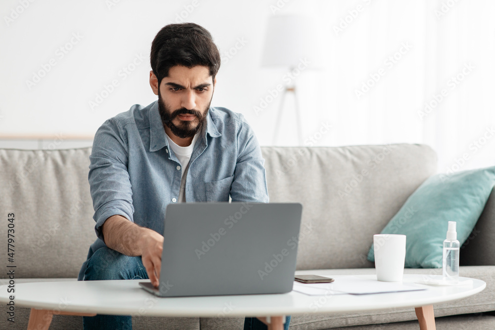 Remote work. Handsome arab guy working on laptop at home office, sitting on sofa and using computer, empty space Remote work. Handsome arab guy working on laptop at home office, sitting on sofa and using computer, empty space