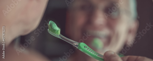 Green toothpaste on toothbrush in hand of senior man looking in mirror