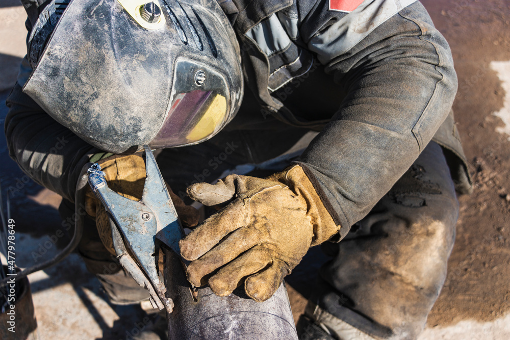 A welder in work clothes is welding a pipe at a construction site. Close-up.