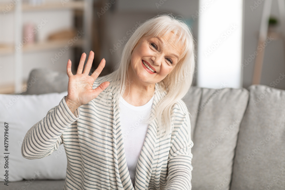 Elderly woman waving at camera and smiling, sitting on sofa