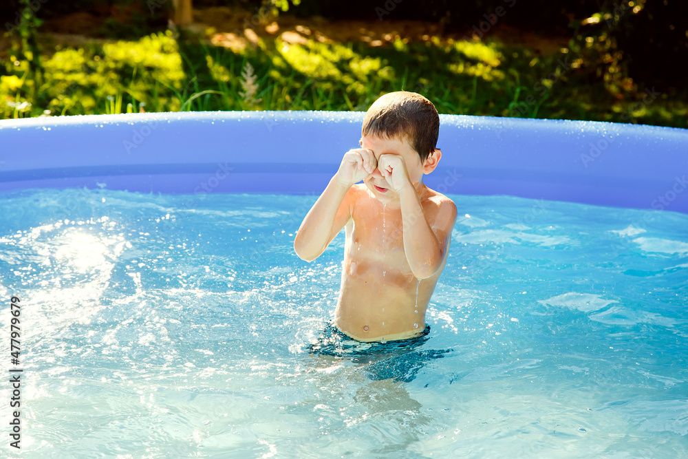 A boy swims in an inflatable pool in the garden on a sunny summer day. He rubs his eyes, from the water that has got in.