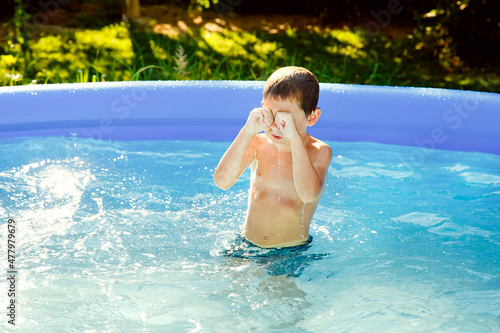A boy swims in an inflatable pool in the garden on a sunny summer day. He rubs his eyes, from the water that has got in.