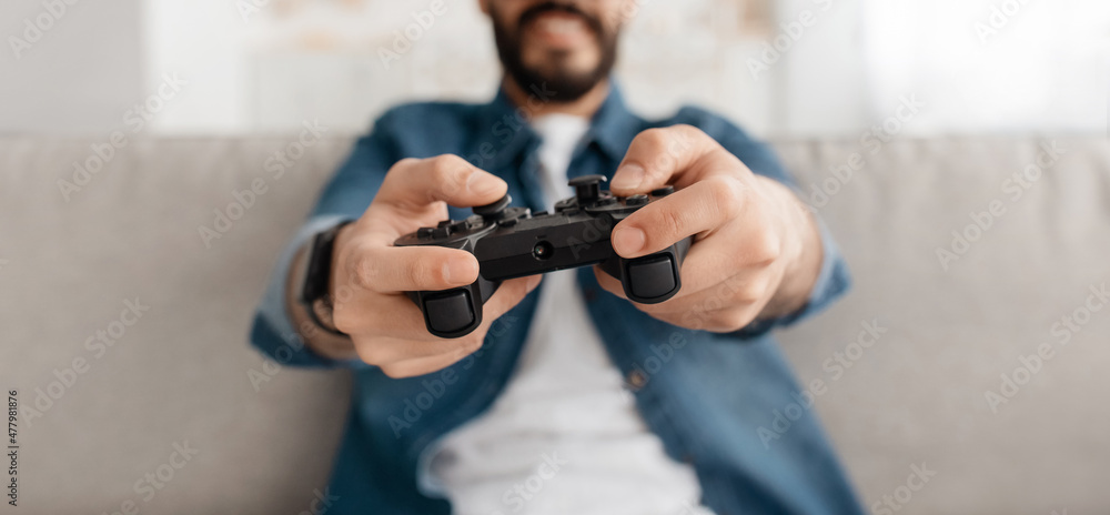 Unrecognizable young guy playing videogames with joystick at home, sitting on sofa, selective focus, crop, panorama